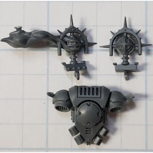 May include: Three gray plastic pieces for a model kit. One piece is a torso with a round center and two side panels. The other two pieces are decorative, one with a round center and the other with a curved shape.