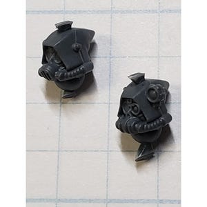 May include: Two unpainted dark gray plastic miniature heads featuring hooded cowls, circular ocular lenses, and integrated respirator masks with ribbed tubing details, designed for tabletop gaming figures.