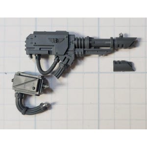May include: A gray plastic miniature weapon component featuring a long barrel with ribbed detailing, a central housing with an embossed winged skull emblem, a separate rectangular power pack, and a flexible cable connector for assembly.