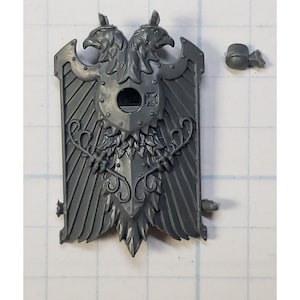 May include: A gray plastic miniature of a double-headed eagle shield with a central round shield. The shield has a raised design of two eagles with wings spread, and a central round shield with a raised design of a circle with a hole in the center. The shield is part of a miniature wargaming set.