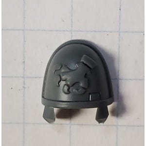 May include: Gray plastic miniature shoulder pad with a raised, detailed design of a stylized scorpion.