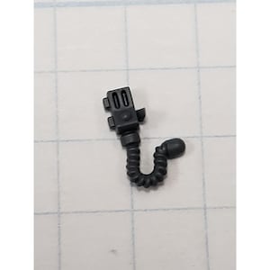 Puede incluir: Un pequeño componente de plástico gris oscuro con una sección superior rectangular con dos aberturas rectangulares. Un tubo flexible y acanalado se extiende hacia abajo, terminando en un extremo redondeado. El objeto está sobre un fondo de cuadrícula.