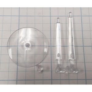 May include: Clear plastic doll stand components. Includes a round base, two support rods, and a small peg. The pieces are transparent and appear to be made of acrylic or a similar material. The components are designed to hold a doll upright.