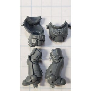 May include: A collection of unpainted, gray plastic miniature parts for a tabletop game. The components include a torso, chest piece with a skull and wing emblem, and two leg pieces. The parts are designed for assembly.