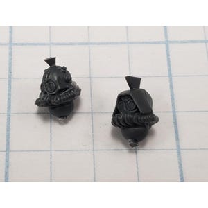 May include: Two unpainted dark gray plastic miniature heads featuring detailed gas masks with circular goggles, breathing apparatuses, and small top-mounted crests. These small gaming components are molded with intricate mechanical textures.