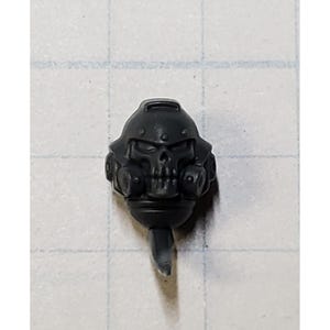 May include: A dark gray miniature helmet with a skull face design. The helmet has a respirator and a small antenna-like piece extending from the bottom. The helmet is likely for a miniature figure or model.