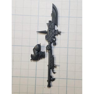 May include: A dark gray plastic miniature weapon with intricate details, including a blade, spear point, and ornate embellishments. The weapon is accompanied by a detached armor piece, all set against a grid background.