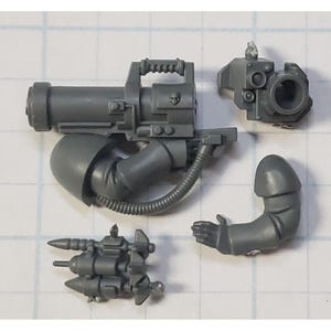 May include: A collection of gray plastic miniature weapon components, including a large gun with a handle, a smaller targeting device, a missile launcher, and an arm with a hand. The pieces are for a miniature model.