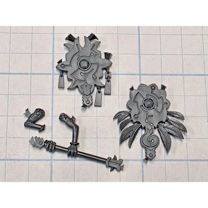 May include: A set of three dark gray plastic model components including two ornate, shield-like decorative pieces with intricate geometric and leaf-like engravings, and a separate rod-shaped piece with two attached hand-like appendages.