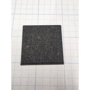 May include: A square black base with a textured, granular surface finish. The object is positioned on a white grid background, providing a clear view of its flat, uniform shape and dark, matte appearance, which is designed for mounting small items.