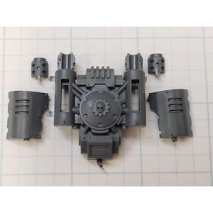 May include: A collection of unpainted, gray plastic miniature wargaming components. The central piece features a circular design with a skull and gear emblem. Additional parts include two side panels, two cylindrical pieces, and two rectangular pieces.