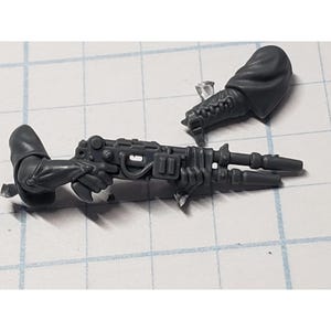 May include: A gray plastic miniature weapon with two arms, a rifle, and a hooded sleeve. The rifle has a detailed design with various mechanical components. The miniature is likely for tabletop gaming or model building.