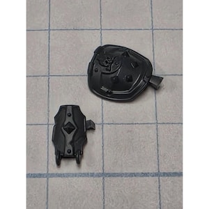 May include: Two dark gray plastic miniature armor components. One piece is a rounded shoulder pauldron featuring a small skull emblem and raised spikes. The second piece is a rectangular forearm bracer with a central diamond-shaped protrusion.
