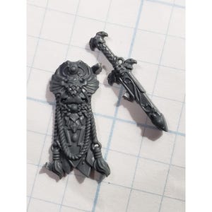 May include: A gray plastic miniature shield and sword. The shield has an ornate design with a central emblem and hanging tassels. The sword features a detailed hilt and blade. The items are likely for a tabletop game or model kit.