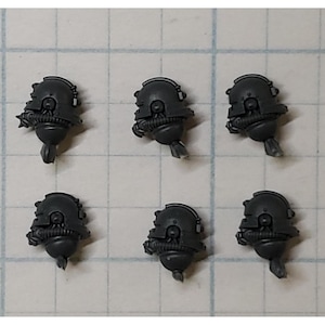 May include: Six dark gray miniature helmet heads, likely for a tabletop game. Each head features a rounded helmet shape with detailed facial features and a lower section with a curved appendage. The items are arranged on a grid.