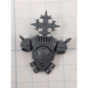 May include: A gray plastic miniature torso with a spiked, cross-shaped ornament on top. The torso has a series of rectangular vents and two rounded shoulder pads with small protrusions. The overall design suggests a fantasy or sci-fi theme.