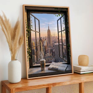 May include: A framed wall art print depicting an open window view of the New York City skyline at sunset, featuring the Empire State Building, a steaming coffee cup, and a newspaper titled The Daily Manhattan resting on the window ledge.