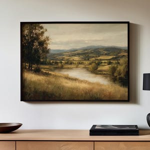 May include: Framed landscape painting depicting a serene river scene with rolling hills and a cloudy sky. The artwork features a palette of muted greens, yellows, and browns, creating a tranquil and naturalistic aesthetic. The painting is displayed in a dark wooden frame.