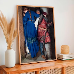 May include: A framed print of a medieval scene. A woman in a long blue dress leans against a stone wall, while a man in armor and a red tunic embraces her. A sword is visible. The artwork is displayed on a wooden table.