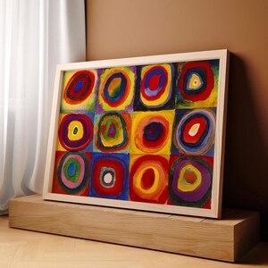 May include: Abstract art print featuring a grid of colorful concentric circles in red, blue, yellow, and green. The artwork is framed in a light wood frame and rests on a wooden block.