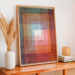 Op de afbeelding: Abstracte geometrische kunstafdruk in een houten lijst. Het kunstwerk toont concentrische vierkanten in tinten rood, oranje, roze, blauw en bruin. Het kunstwerk wordt getoond op een houten kast met een vaas met gedroogde planten.