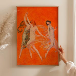 May include: A framed rectangular wall art print featuring three figures in flowing, translucent garments dancing against a vibrant, solid orange background. The figures are depicted in motion with arms raised and fabric trailing behind them in a dynamic composition.
