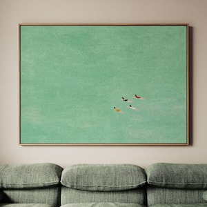 May include: A framed rectangular wall art print featuring a large expanse of textured mint green water. Four small figures are depicted swimming in the water, each wearing a different colored swimsuit including black, red, yellow, and white.
