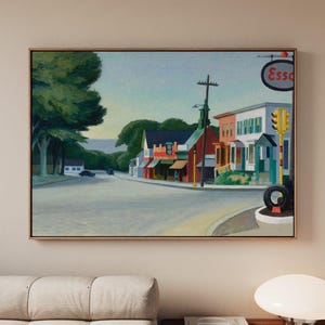 May include: A framed landscape painting depicting a quiet street corner with colorful buildings, a tall utility pole, and an Esso sign. The scene features a large green tree on the left, a traffic light on the right, and a clear sky above the road.