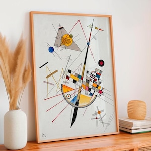 May include: Abstract art print in a light wood frame. The artwork features geometric shapes, lines, and colors including yellow, red, blue, and black. The print is displayed on a wooden surface, with a vase of dried plants and a small woven basket in the background.