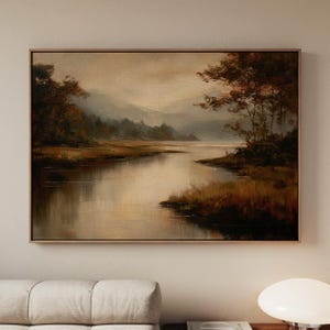 May include: A framed landscape painting depicts a serene river scene. The artwork features a muted color palette of browns, greens, and golds, with a reflective river and trees along the banks. The painting is encased in a thin, gold-toned frame.