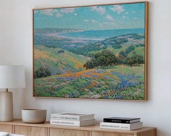May include: A rectangular landscape painting in a light wood frame depicting a coastal valley filled with orange and blue wildflowers, rolling green hills, and a distant blue ocean under a bright sky with scattered white clouds.