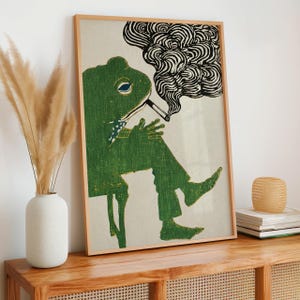 May include: A framed art print featuring a green frog smoking a cigarette. The frog is wearing a blue and white patterned bow tie and is seated. The smoke is black and swirly. The art print is in a wooden frame.