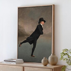 May include: A framed art print depicting a person in a black coat, breeches, and a top hat ice skating on a frozen lake. The figure is captured in a gliding pose against a muted, cloudy sky and a landscape background with soft, neutral tones.