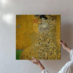 May include: Square canvas print of a Gustav Klimt painting. The artwork features a woman in a long, ornate gold dress with intricate patterns. The background is a blend of gold and yellow hues, with a touch of green.