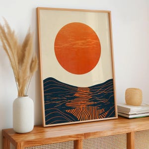 May include: A framed wall art print featuring a large, solid orange circle representing a sun positioned above dark blue, wavy horizontal lines that create a textured landscape effect with a central orange reflection on a cream background.