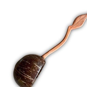 Może przedstawiać: Ręcznie wykonana drewniana łyżka z miseczką z łupiny kokosa. Uchwyt wygina się wdzięcznie, kończąc się na wzorze w kształcie liścia. Łyżka wykonana jest z jasnobrązowego drewna, a miska ma kolor ciemnobrązowy.