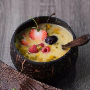 May include: A black coconut bowl filled with a yellow smoothie topped with fresh fruit, including strawberries, raspberries, and a cherry. A wooden spoon rests on the rim of the bowl.