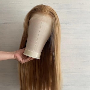 Könnte beinhalten: Eine lange, hellbraune Perücke mit natürlichem Haaransatz wird auf einem Schaufensterkopf präsentiert. Die Perücke hat glattes, fließendes Haar. Der Hintergrund ist weiß und hellblau, und die Perücke wird von einer Hand gehalten.