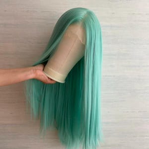 May include: A long, straight wig in a vibrant teal color. The wig is styled on a mannequin head, showcasing its length and the natural-looking hairline. The hair is a solid color, with a slight sheen.