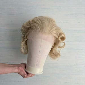 May include: A blonde wig with styled curls is displayed on a mannequin head. The wig has a natural-looking hairline. The mannequin head is held by a person's hand against a light blue and white background.