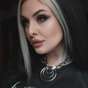 May include: A woman with black and white hair is wearing a black t-shirt with white text that says "behave". She is wearing a silver chain choker with three large silver rings.