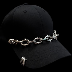 Può includere: Un cappellino da baseball nero a tinta unita con una catena in metallo color argento sul pannello frontale. La catena è composta da maglie allungate e appuntite. Un piccolo anello in metallo argentato a tre punte è fissato sulla visiera.
