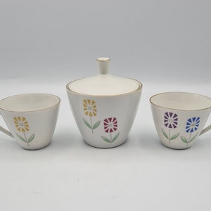 Può includere: Una zuccheriera in ceramica bianca con coperchio e due tazze da tè abbinate. La zuccheriera e le tazze da tè sono decorate con un motivo floreale con fiori gialli, rossi, blu e verdi.