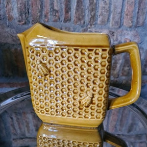 May include: A yellow ceramic pitcher with a honeycomb pattern and bee designs. The pitcher has a handle and a spout.