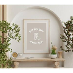 Puede incluir: Una impresión enmarcada beige con un gráfico blanco de botas de vaquero y el texto "Gold Rush Ranch A Photo Finish".