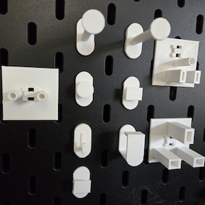 May include: A collection of white plastic pegboard hooks and organisers. The hooks are various shapes and sizes, including a single hook, a double hook, and a triple hook. The organisers are also various shapes and sizes, including a small square organiser, a rectangular organiser, and a larger square organiser.