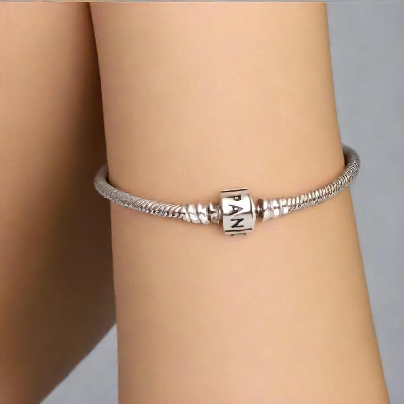 K&ouml;nnte beinhalten: Ein silbernes Armband mit Schlangenkettenmuster. Das Armband hat einen quadratischen Anh&auml;nger mit den Buchstaben "PAN" in Schwarz. Das Armband wird am Arm einer Person getragen.