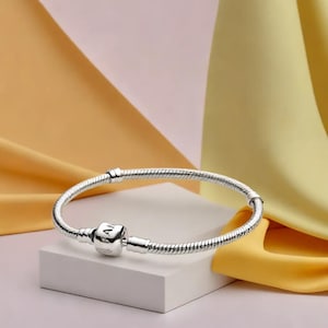 May include: A silver bracelet with a snake chain design. The bracelet has a square charm with engraved text and a clasp. The jewellery is displayed on a white block against a backdrop of yellow and pink fabric.