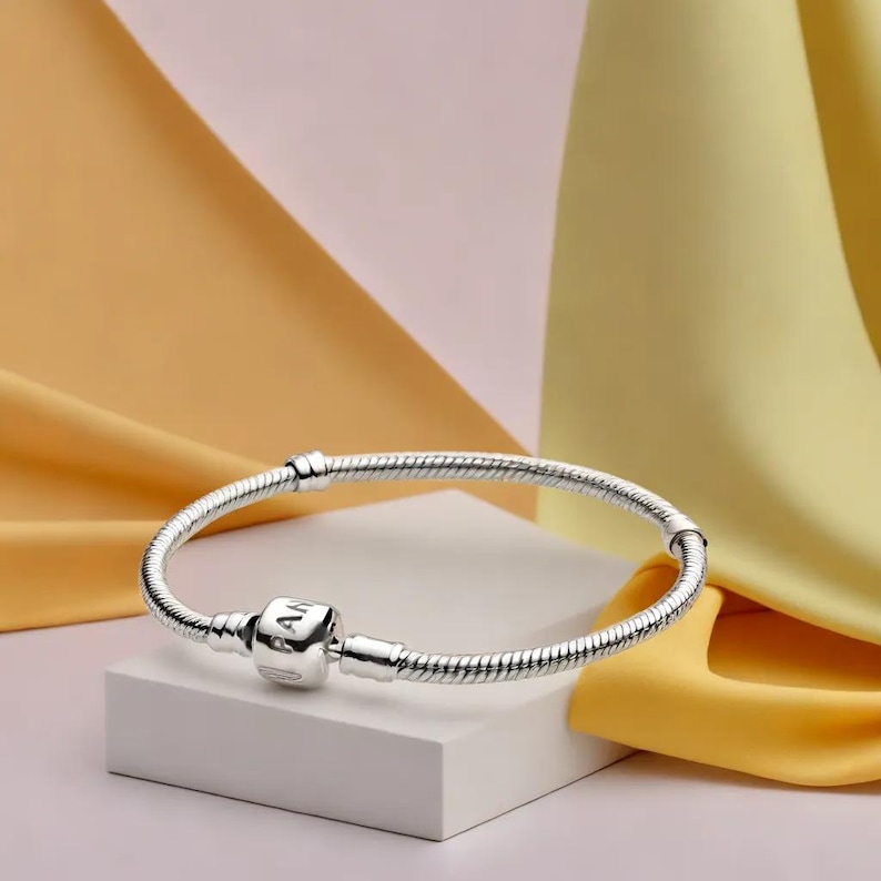 K&ouml;nnte beinhalten: Ein silbernes Armband mit Schlangenketten-Design. Das Armband hat einen quadratischen Anh&auml;nger mit eingraviertem Text und einen Verschluss. Der Schmuck wird auf einem wei&szlig;en Block vor einem Hintergrund aus gelbem und rosa Stoff pr&auml;sentiert.