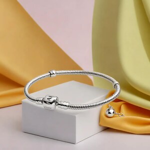 K&ouml;nnte beinhalten: Ein silbernes Armband mit Schlangenkettendesign und einem quadratischen Verschluss mit den Buchstaben "AM". Ein kleiner Charm mit einer silbernen Kugel h&auml;ngt am Armband. Das Armband wird auf einem wei&szlig;en W&uuml;rfel vor farbigem Hintergrund pr&auml;sentiert.